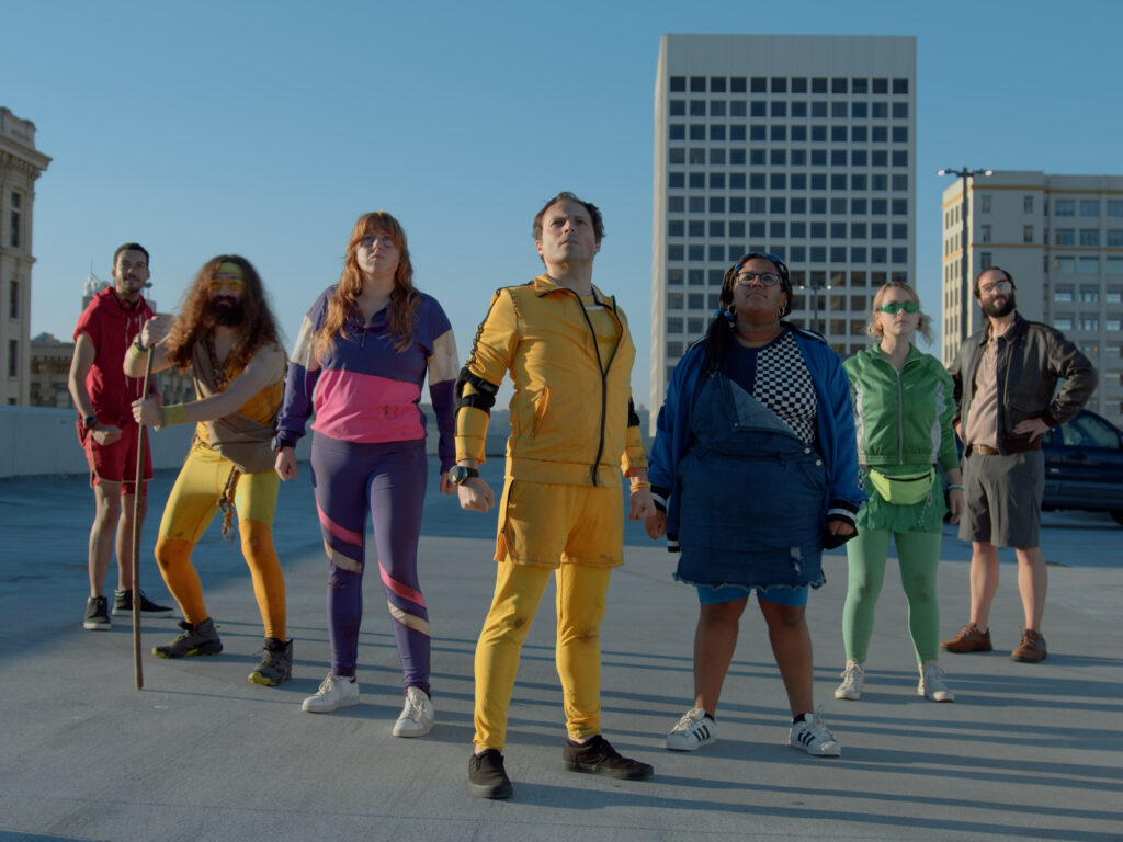 Colorfully clad actors stand on a rooftop in V formation